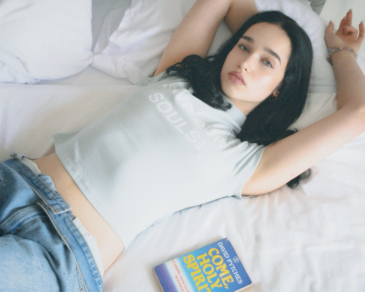 Model lying on white bedding in a pale SOS crop tee, with a book nearby.