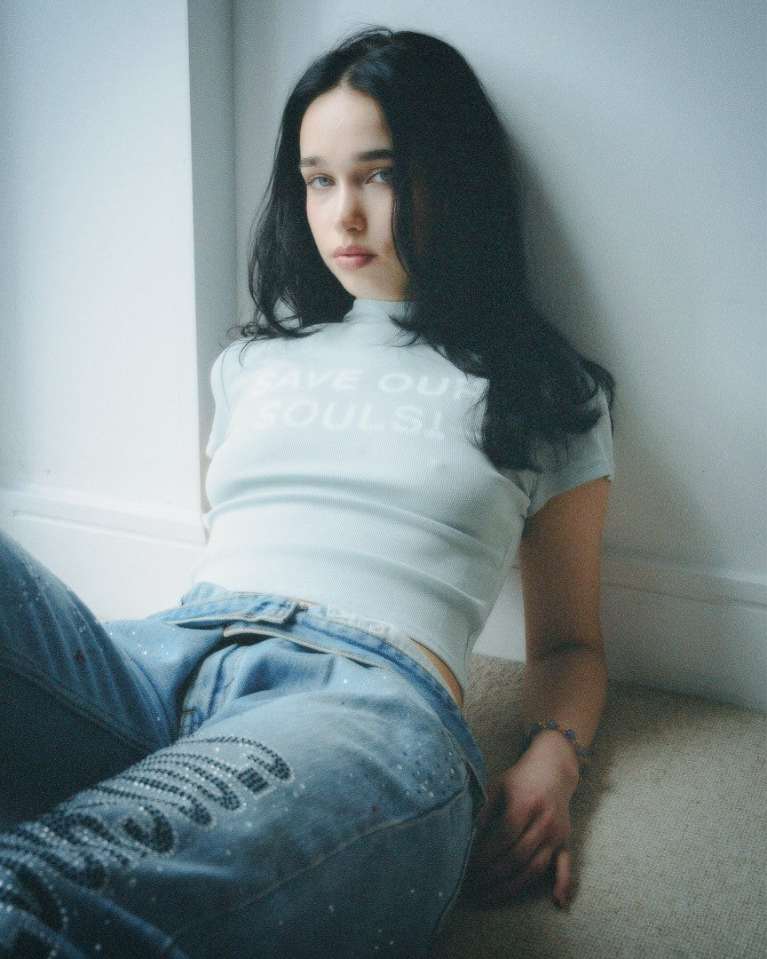 Model seated by a window in a pale SOS crop tee and embellished jeans.