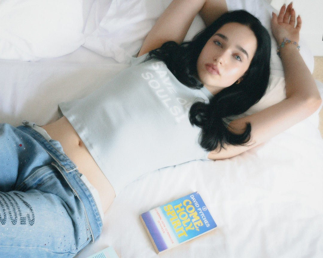 Model reclining on a bed in a pale SOS crop tee with books beside her.