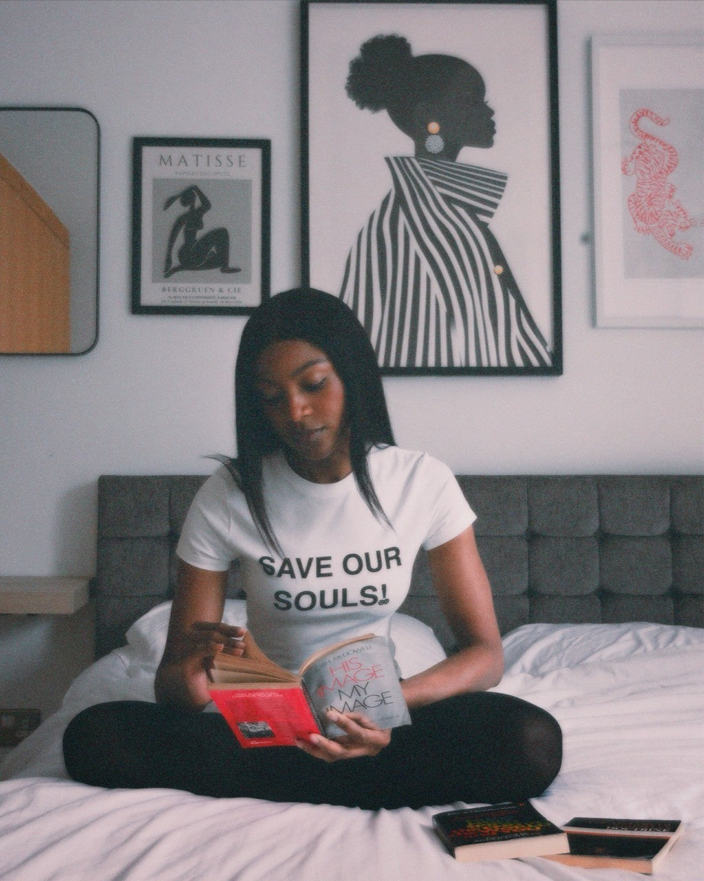 Model sitting cross-legged on a bed and reading in a white SOS tee beneath framed prints.