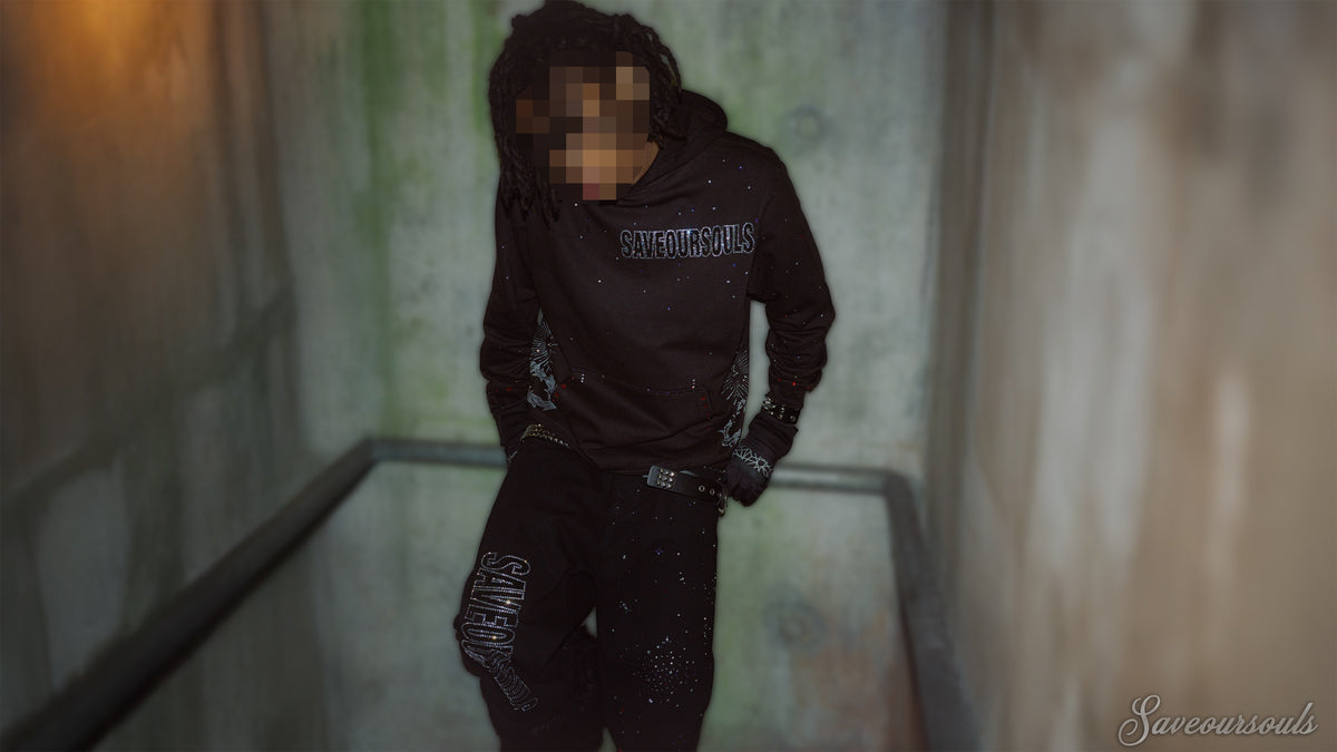 Model wearing a black SOS rhinestone hoodie and dark jeans in a concrete corridor.