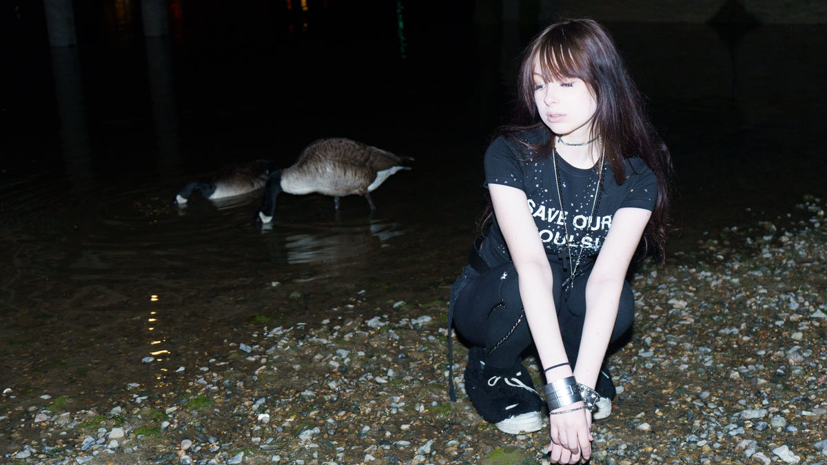 Model crouching beside water at night in a black SOS T-shirt.