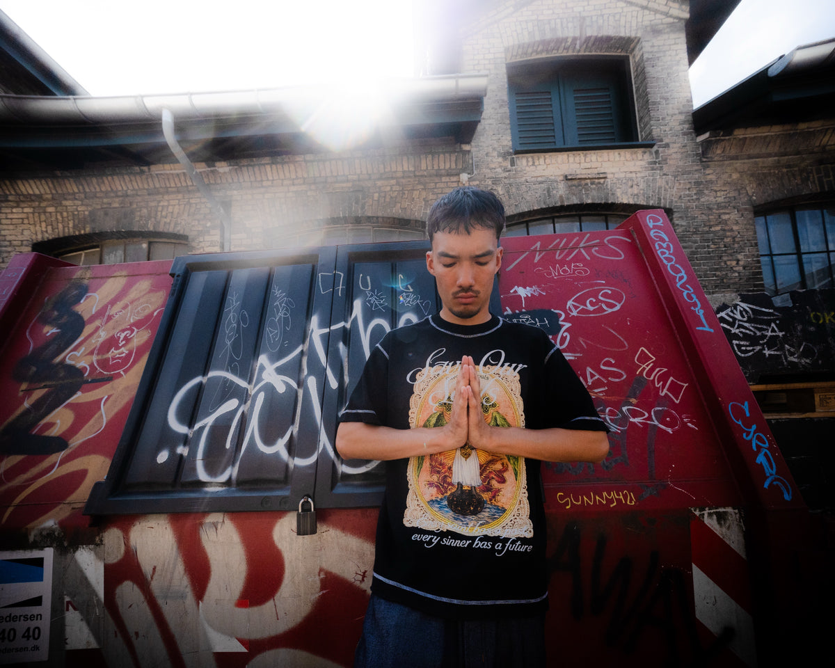 Model wearing a black Angel graphic T-shirt with hands together in a graffiti-lined alley.