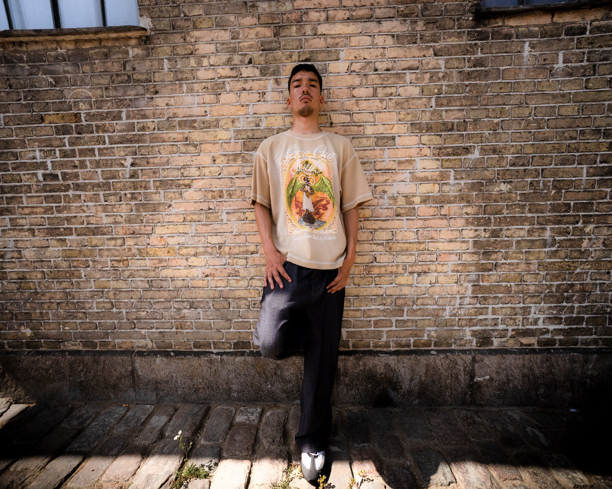 Model standing against a brick wall in a beige Angel graphic T-shirt.