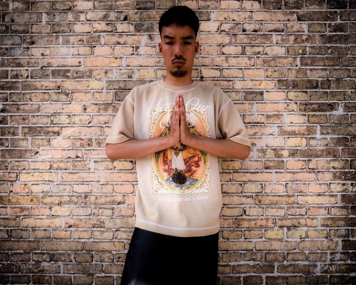 Model wearing a beige Angel graphic T-shirt with hands together against a brick wall.
