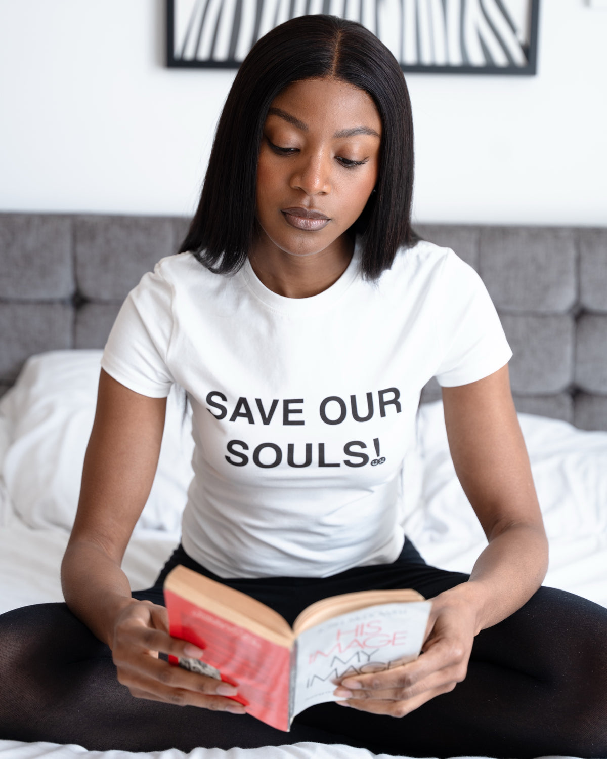 Model reading a book on a white bed while wearing a white SAVE OUR SOULS crop T-shirt.
