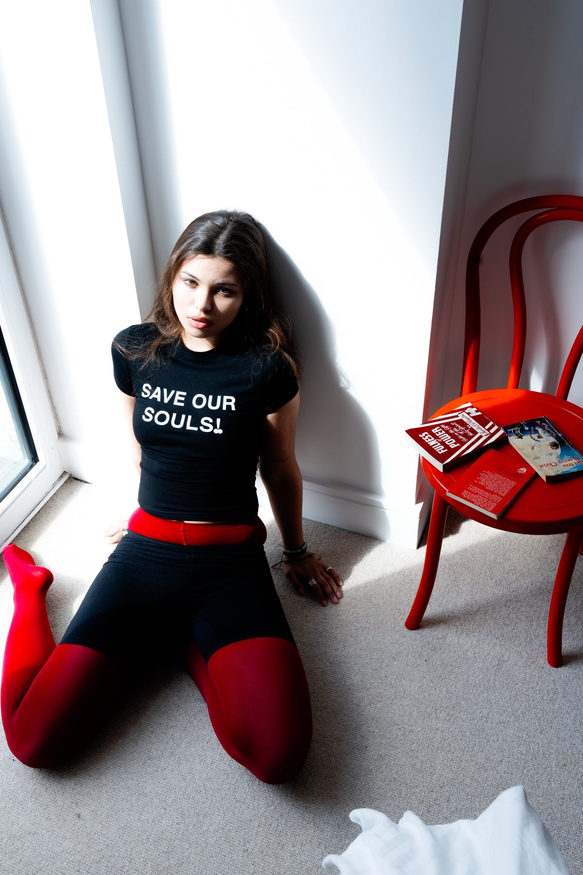 Model wearing a black SAVE OUR SOULS crop T-shirt beside a window and red chair.