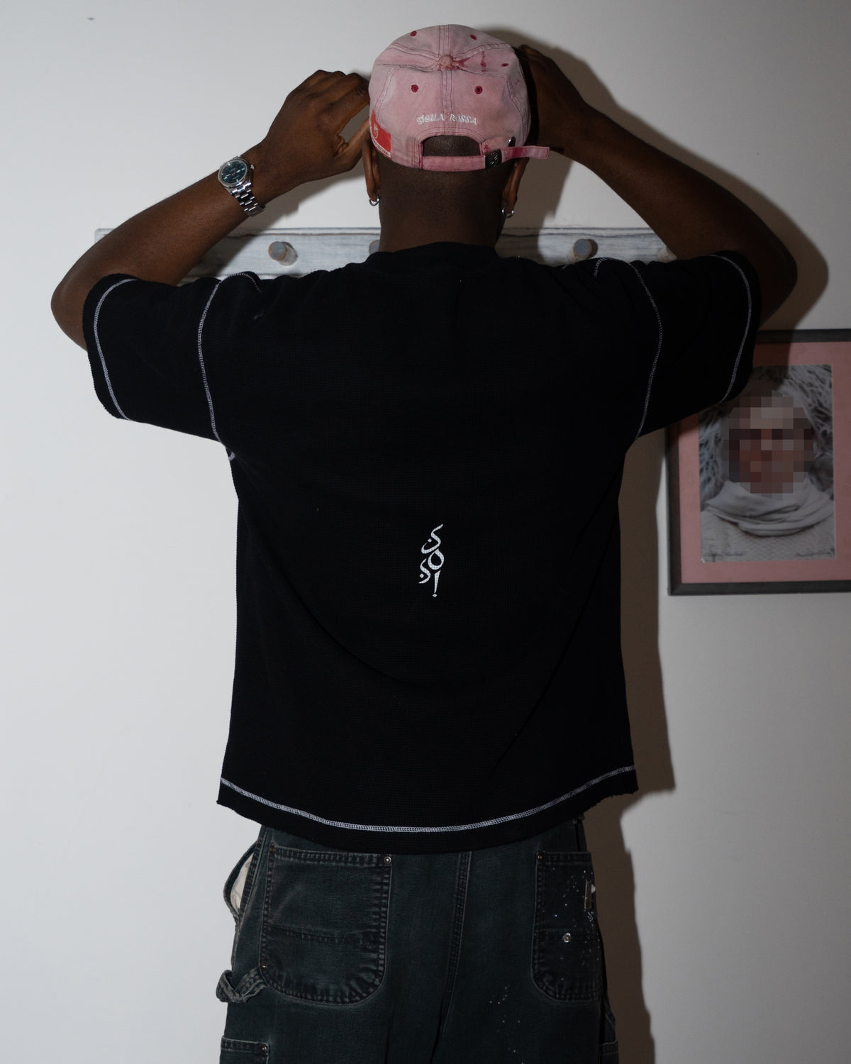 Back view of a black T-shirt with a small SOS emblem, worn with a pink cap.